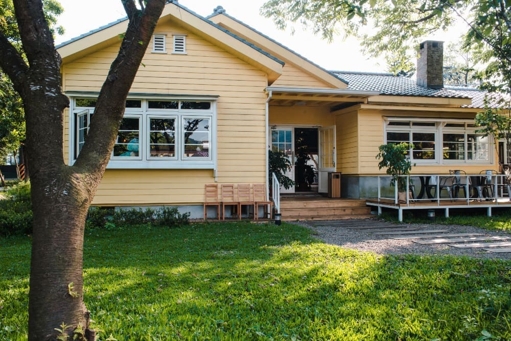 Bright yellow house with a porch and green lawn, surrounded by trees.
